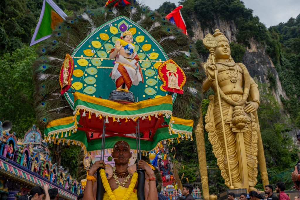 A devotee carries an ornately decorated kavadi in front of the towering golden statue of Lord Murugan at Batu Caves, marking the culmination of the Thaipusam pilgrimage.